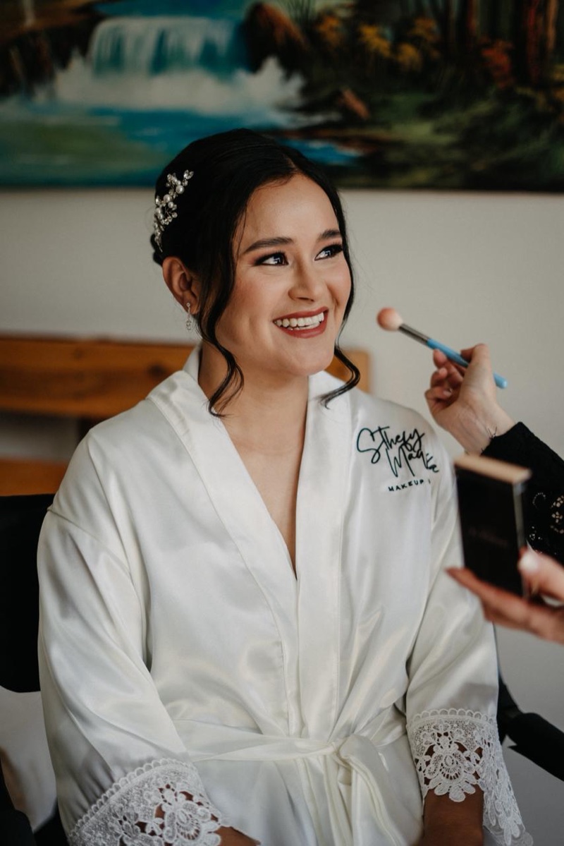 Novia sonriendo en bata blanca mientras le aplican rubor durante su arreglo